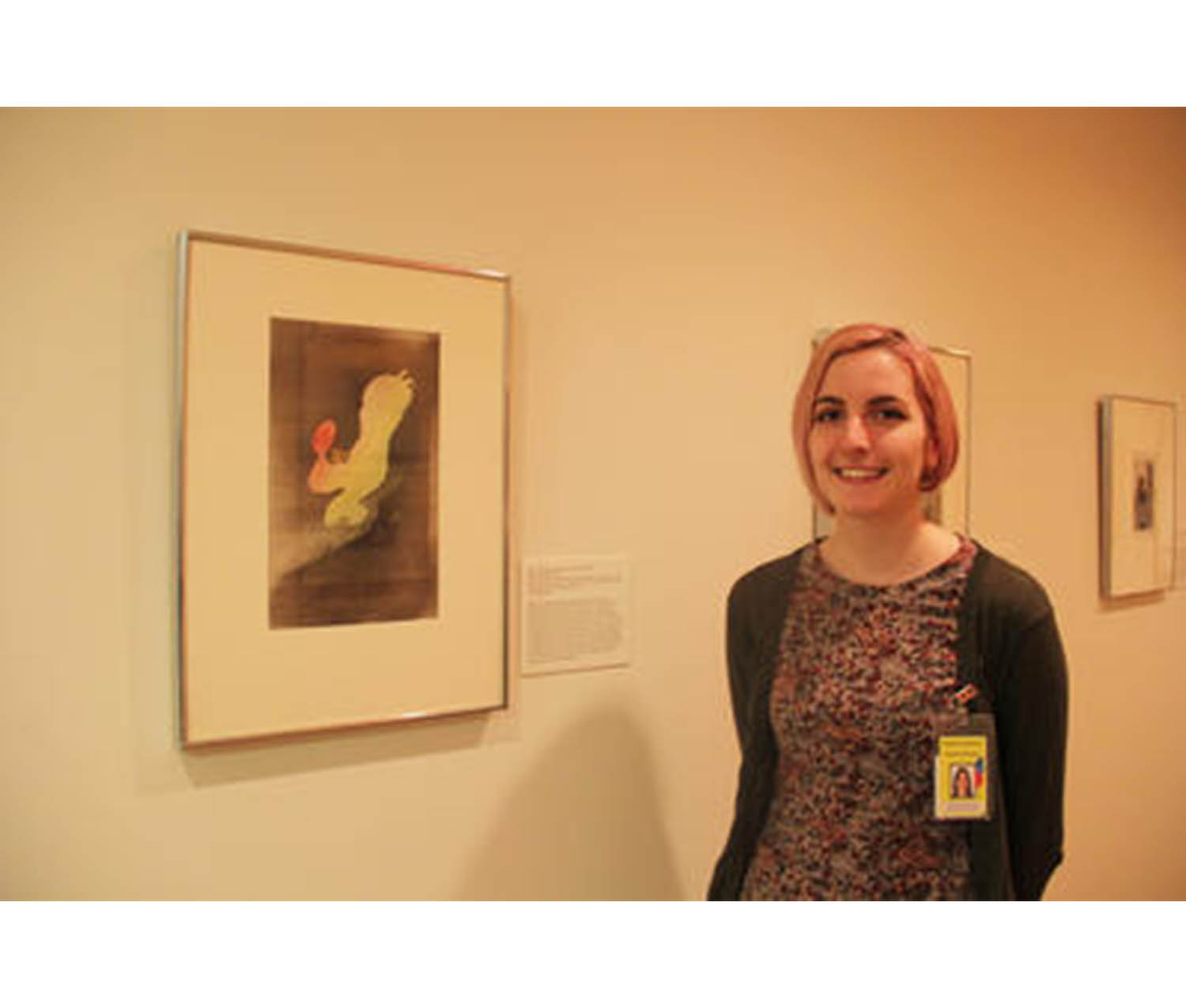 girl wearing a dress and a cardigan with a yellow ID tag clipped onto it, standing next to a framed print and smiling