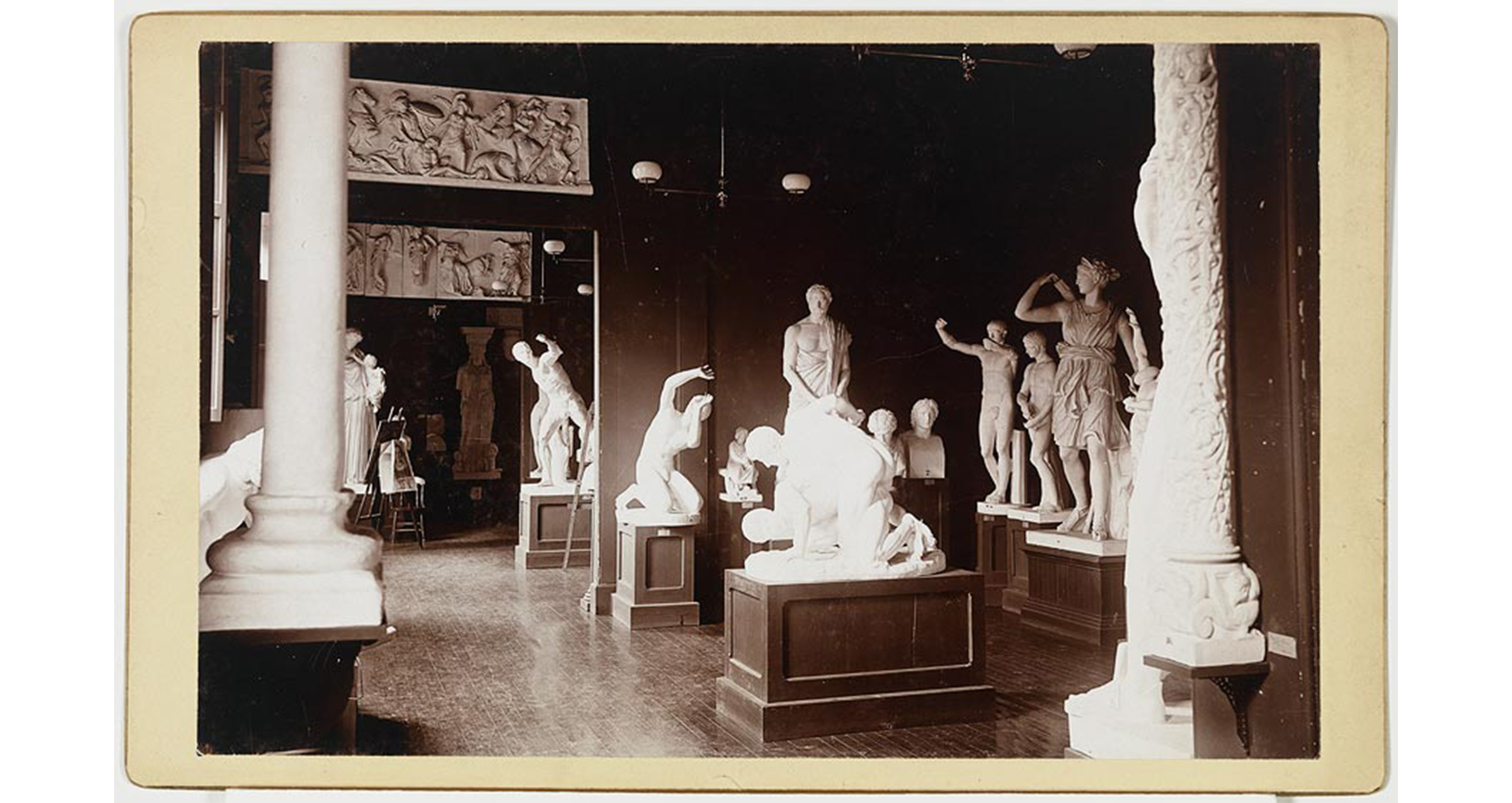 interior, building with pedestals holding plaster casts of classical sculpture and architectural elements, wood floor, hanging gas lighting, two doorways with plaster cast friezes over them