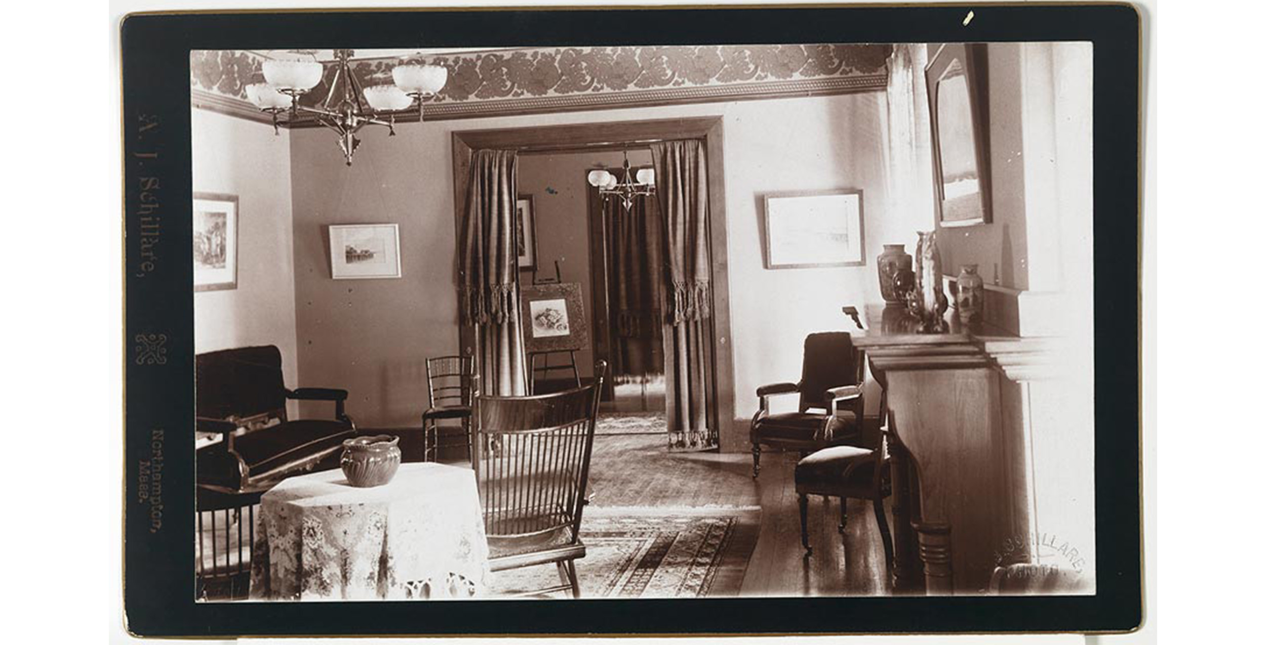 interior, mantle and two padded chairs on right, cloth covered table, rocker, side chair and padded couch opposite, framed pictures on wall, oil ceiling lamp over table, oriental on floor, curtains at large opening to distant hall which has framed picture on easel, one on wall, hanging oil lamp, oriental rug on floor and doorway to another room with closed curtain
