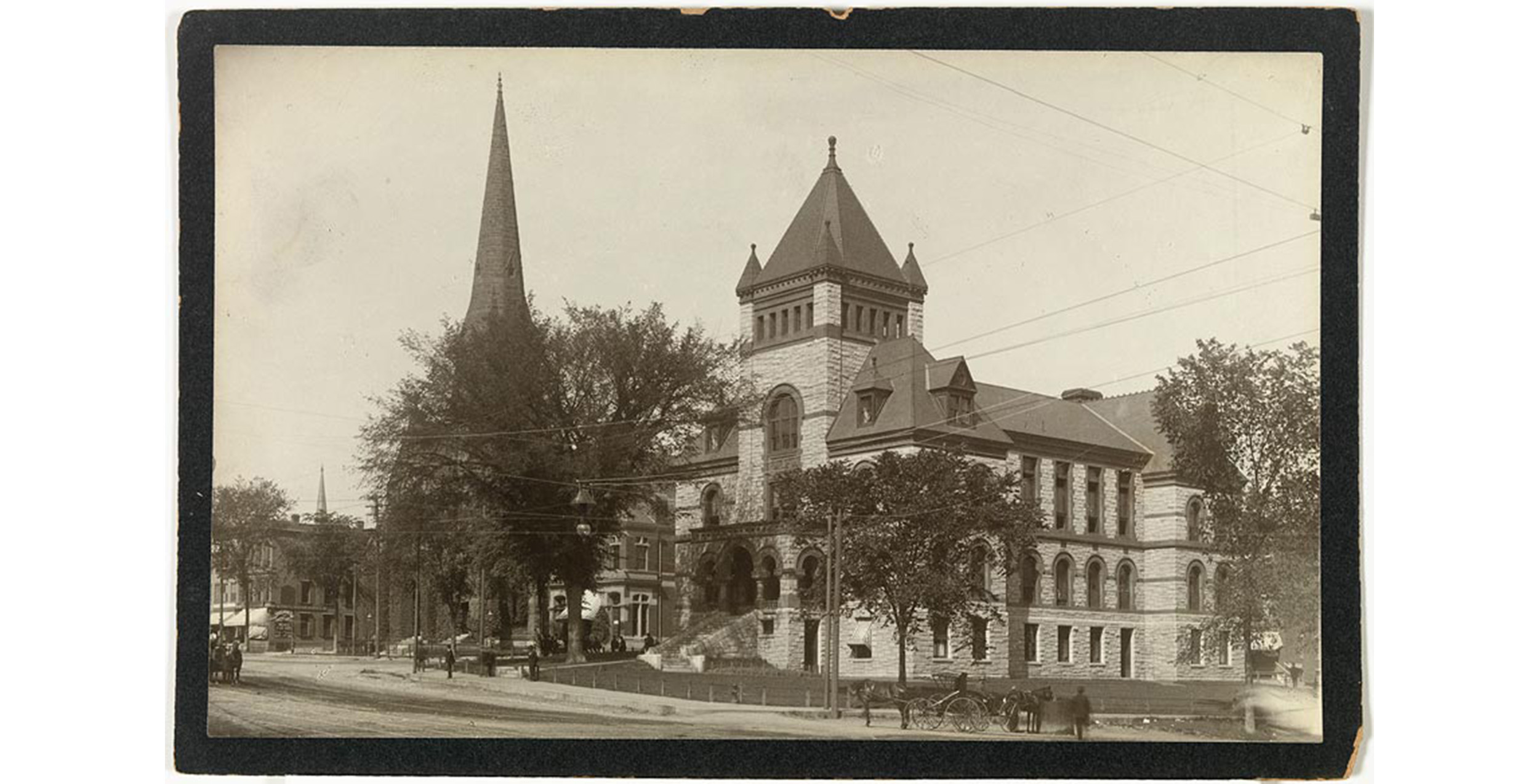 exterior, street curving around large stone building with lawn and trees, visible through trees is church spire, people and horse drawn carriages on street