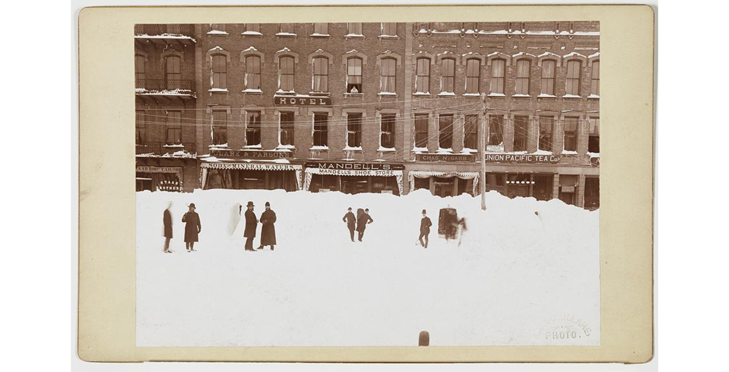 exterior, winter, city street under deep snow with attached brick buildings and eight scattered people standing in snow, signs on buildings include: HOTEL, AAB BROS TAILORS, CLARK & PARSONS, SODA and MINERAL WATERS, MANDELL'S SHOE STORE, CHA'S AND GABB, and UNION PACIFIC TEA Co.