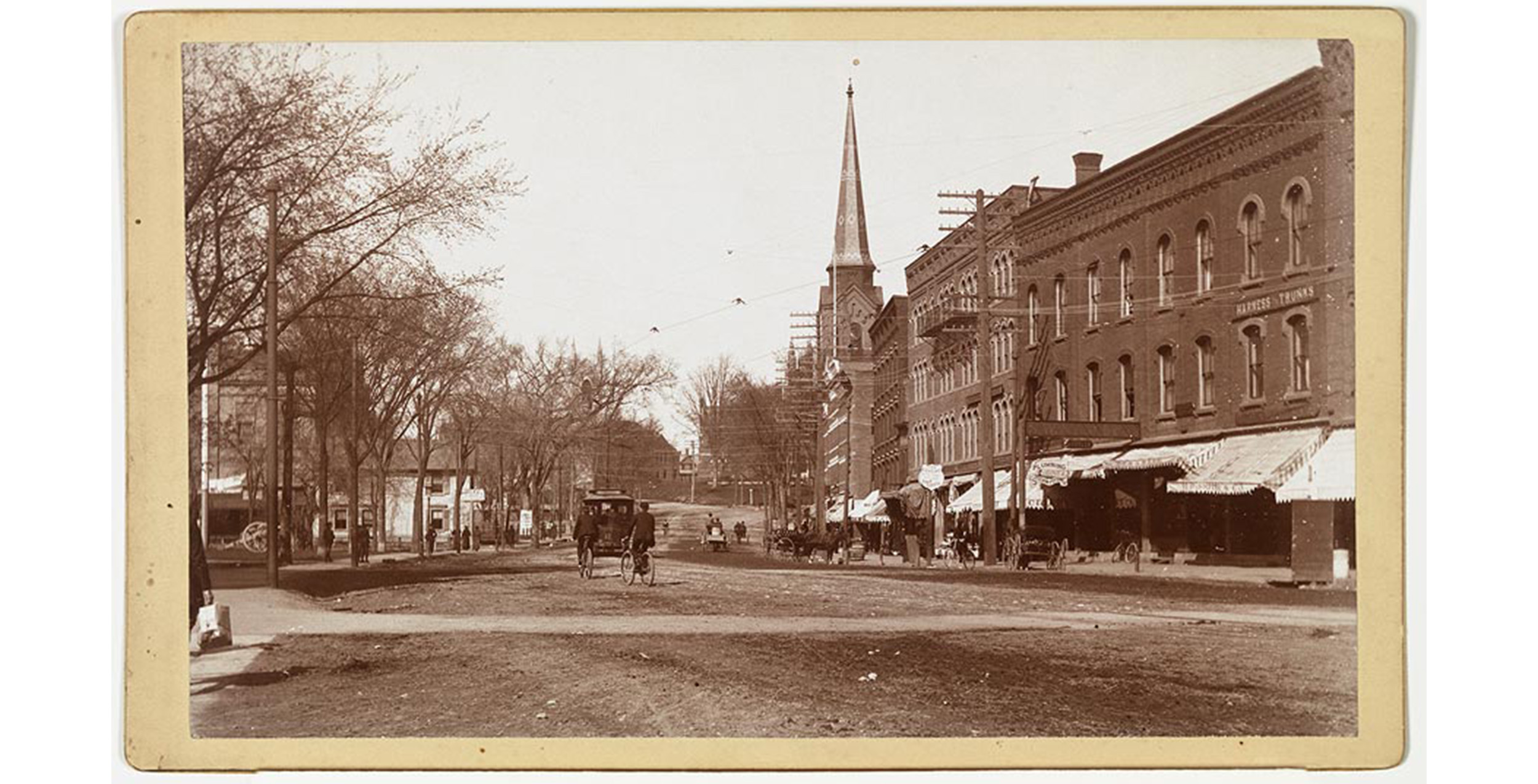 exterior, main street with dirt street, horse drawn carriages and bicyclists on street, brick city block on right with HARNESS TRUNKS sign, awnings covering sidewalk, trees lining street on left, people on sidewalk, telephone poles and wires lining streets, distant view of parts of Smith College buildings