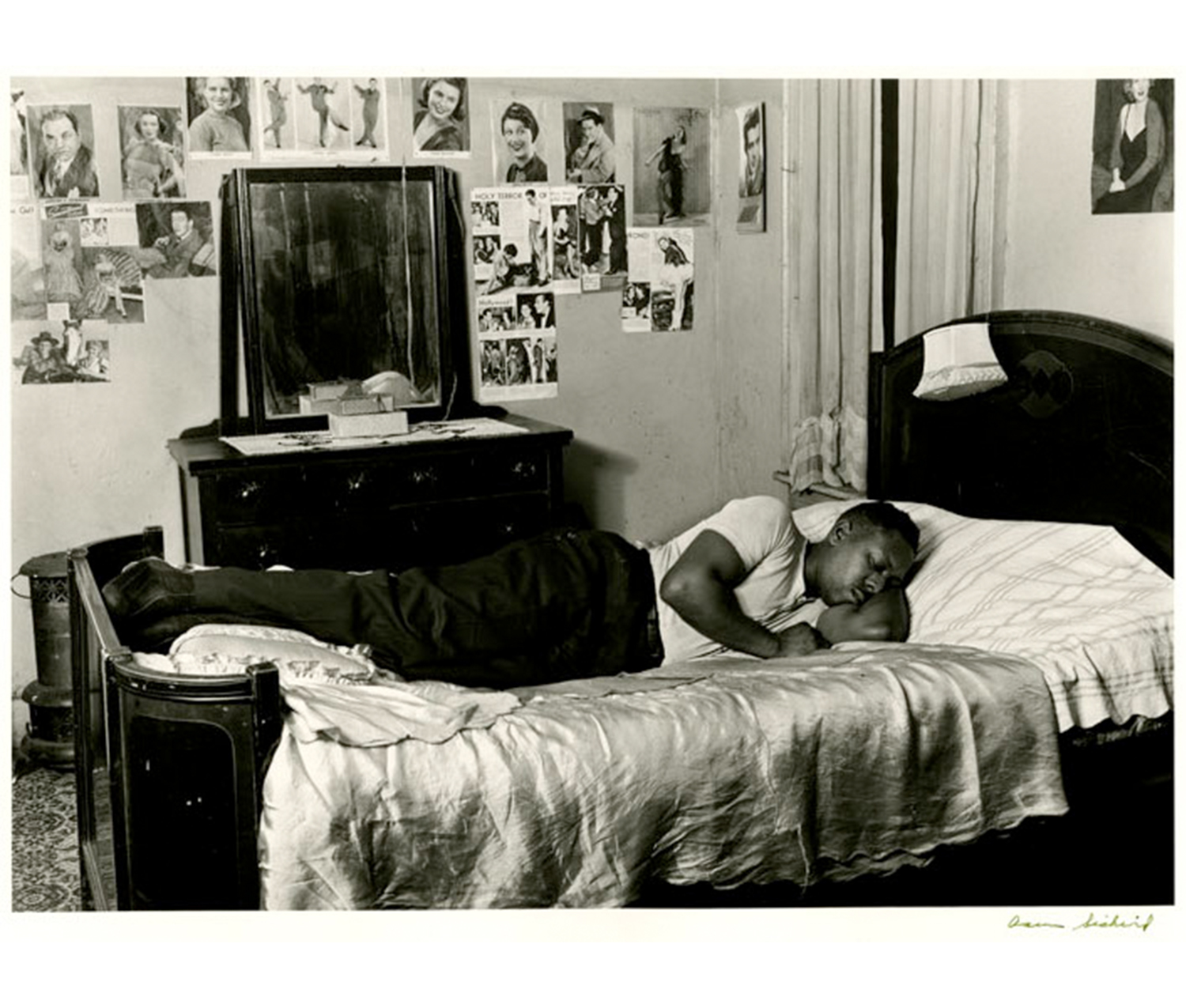 man lying on a bed, turning toward the camera; in the background, a chest of drawers, a mirror, and photographs taped to the wall