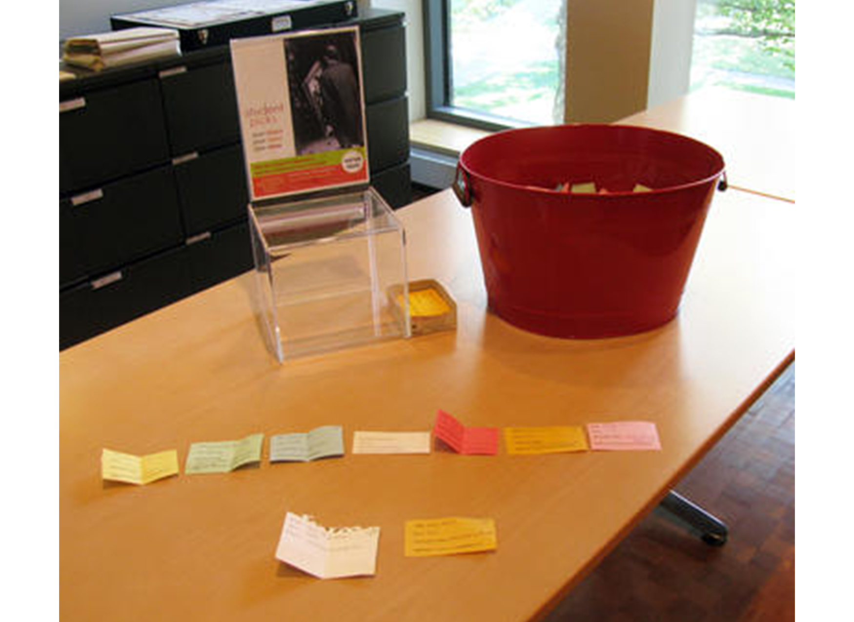 wooden table with a sign propped up on it, a red bucket full of colorful slips of paper, and nine slips of paper arranged in two rows