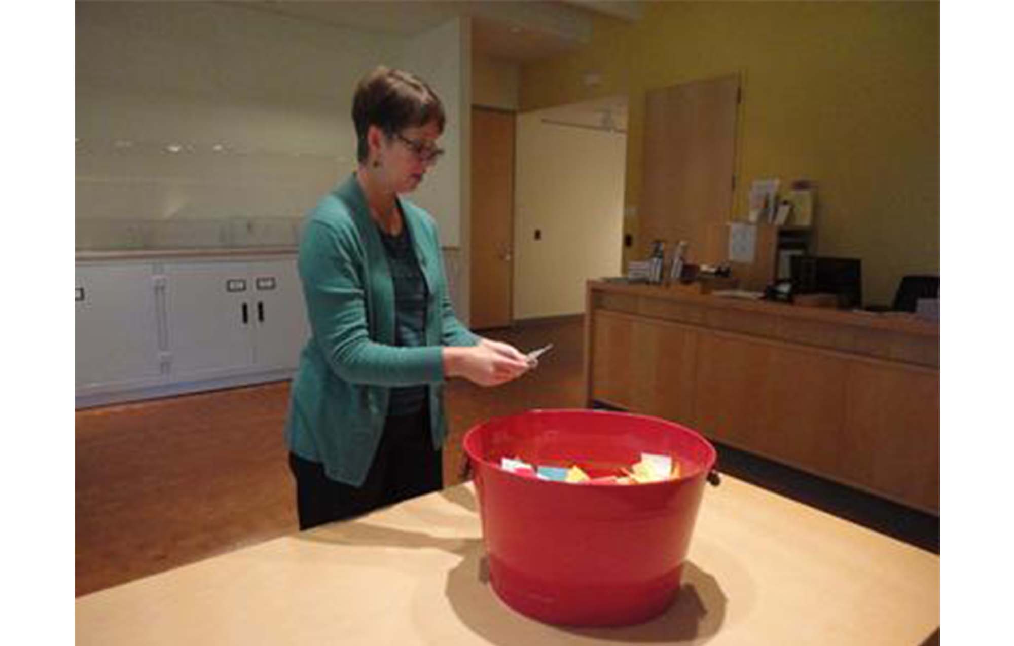 woman stands in front of a table, holding a slip of paper that she has just pulled out of a large red bucket with many slips of paper inside of it
