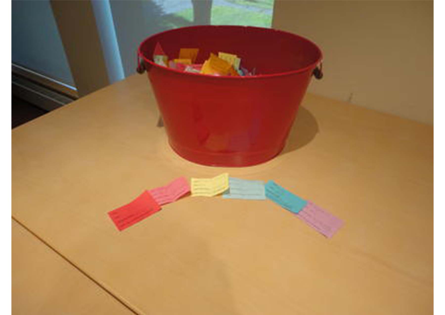 large red bucket with multicolored slips of paper inside of it, sitting on a wooden table. six slips of paper arranged on the table in an arc in rainbow order