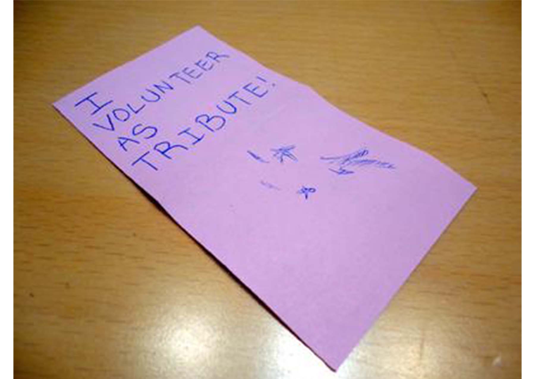 purple slip of paper with "I VOLUNTEER AS TRIBUTE!" written on it, sitting on a wooden tabletop