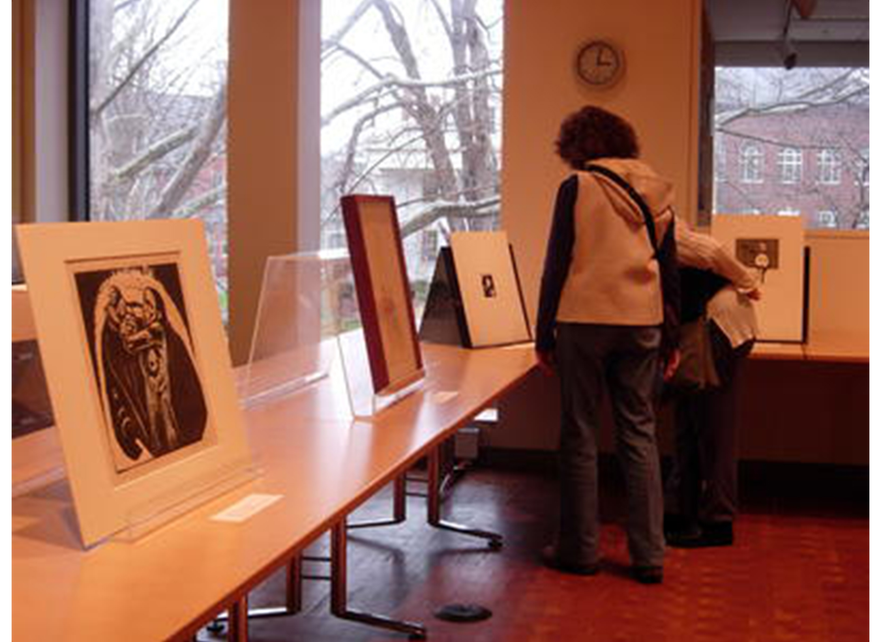 room with tables with printed propped up on them; two people stand in the corner looking closely at one of the prints with their backs to the viewer