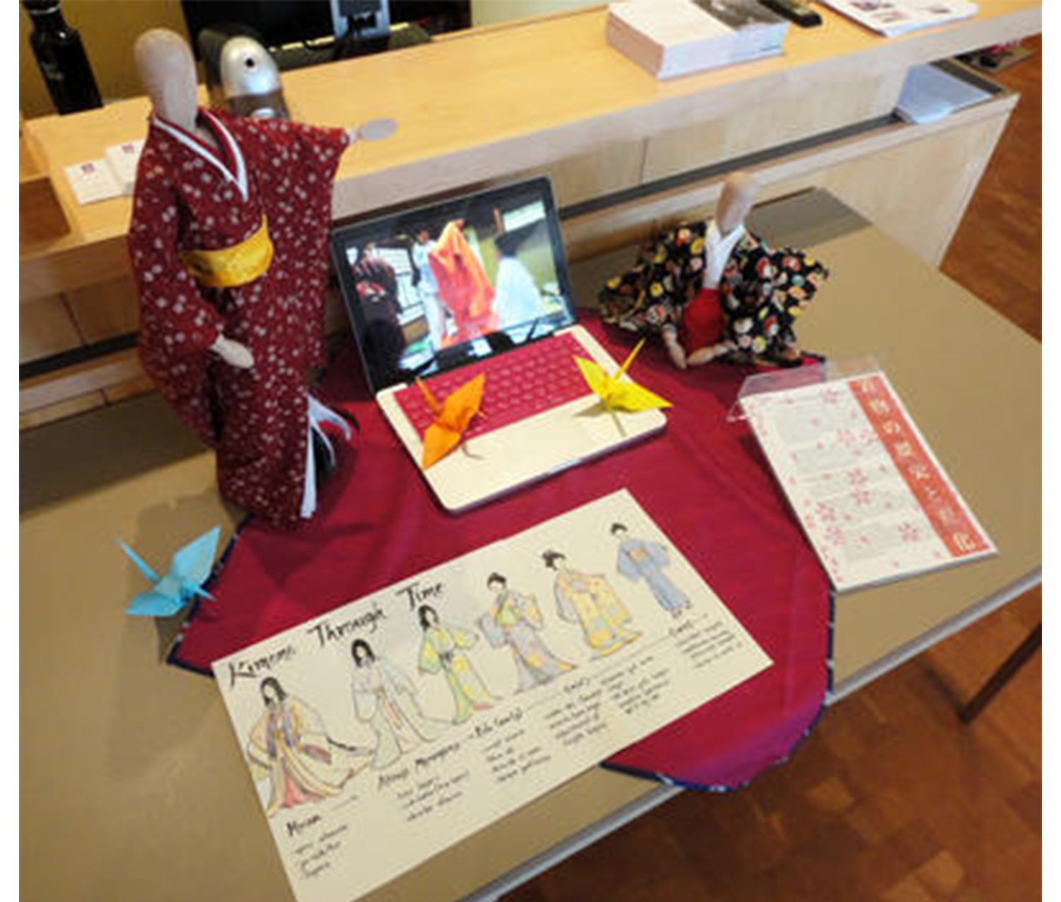 table with a drawing on it, "Kimonos Through Time" written on it with drawings of women wearing kimonos below. also on the table are paper cranes, a laptop with its screen propped up, and two small mannequins wearing kimonos