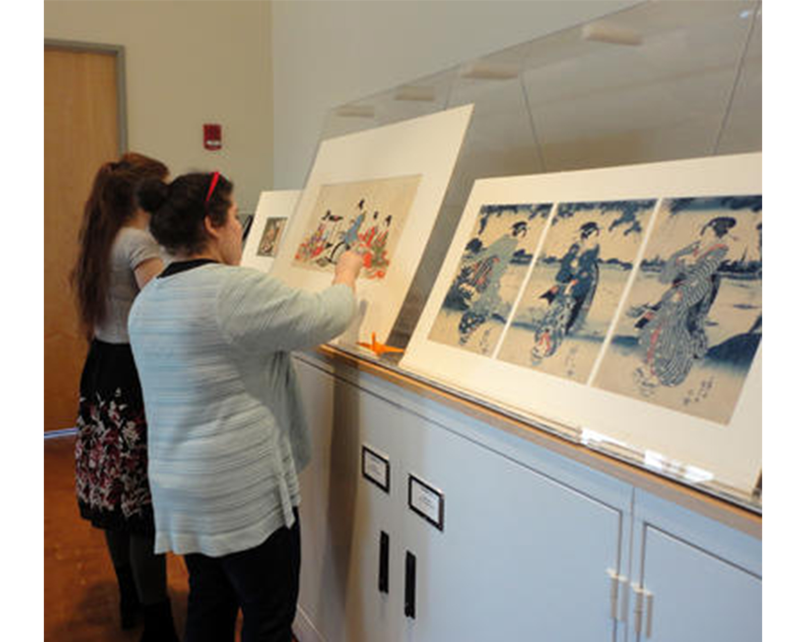 two girls stand in front of prints propped up on a gray cabinet, examining the prints closely and pointing to them