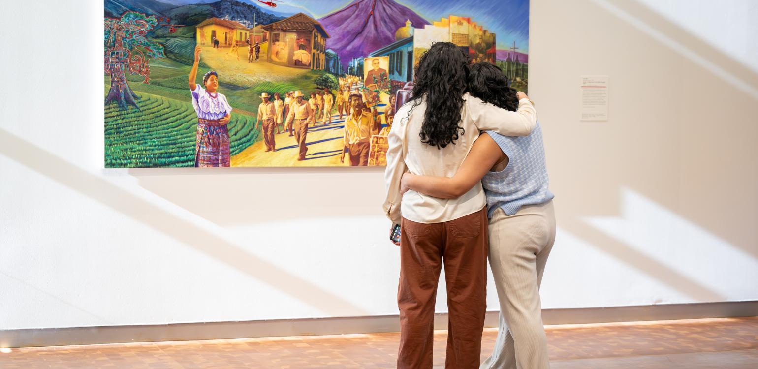 Two people with arms around each other looking at a paitning of people dressed in colorful clothes walking along a road