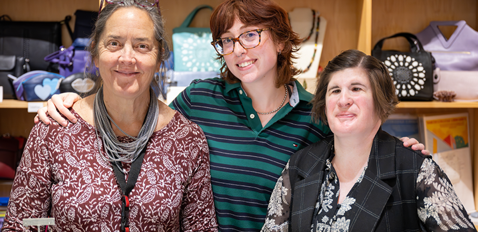 Three people standing together for a photograph with purses on shelves in the background