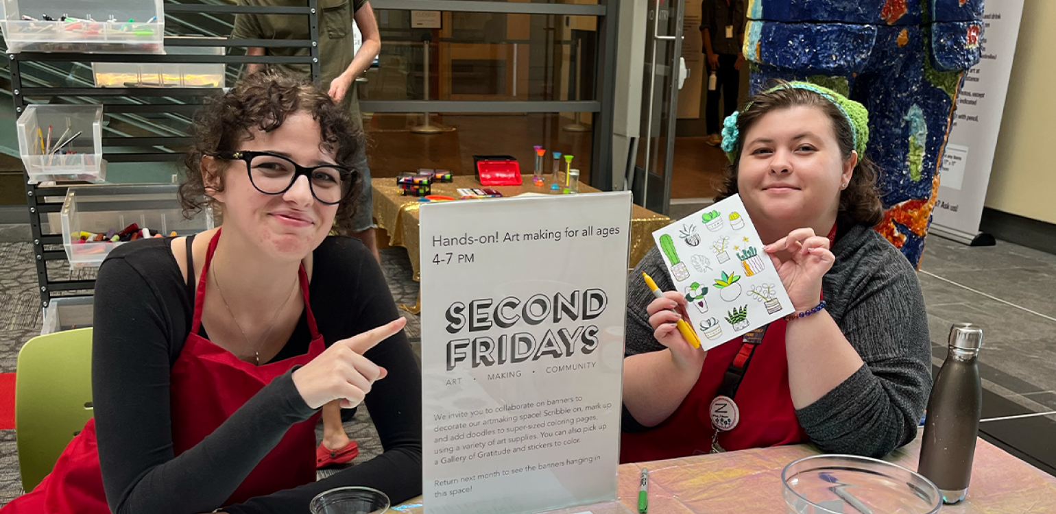 Two people sitting at a table, one pointing to a sign that reads “Second Friday"