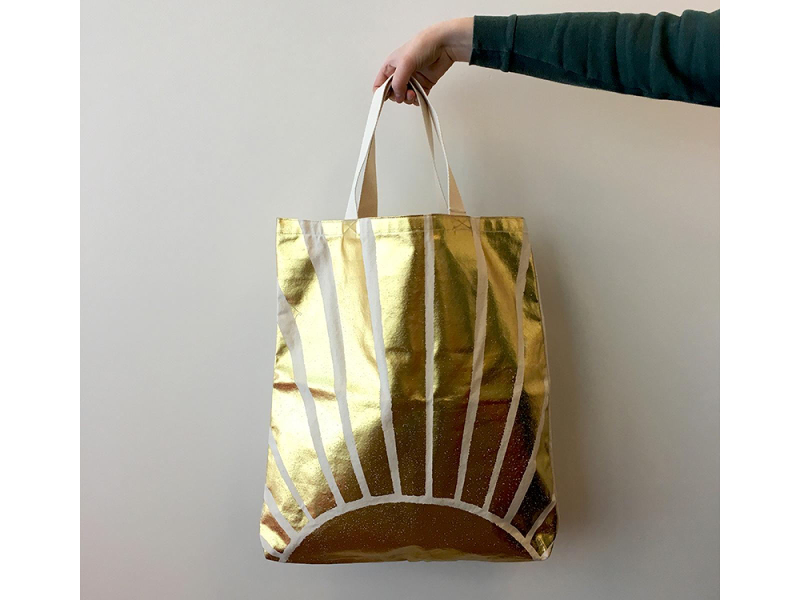 A hand holding a bag, with a golden sun on it, by a strap handle.