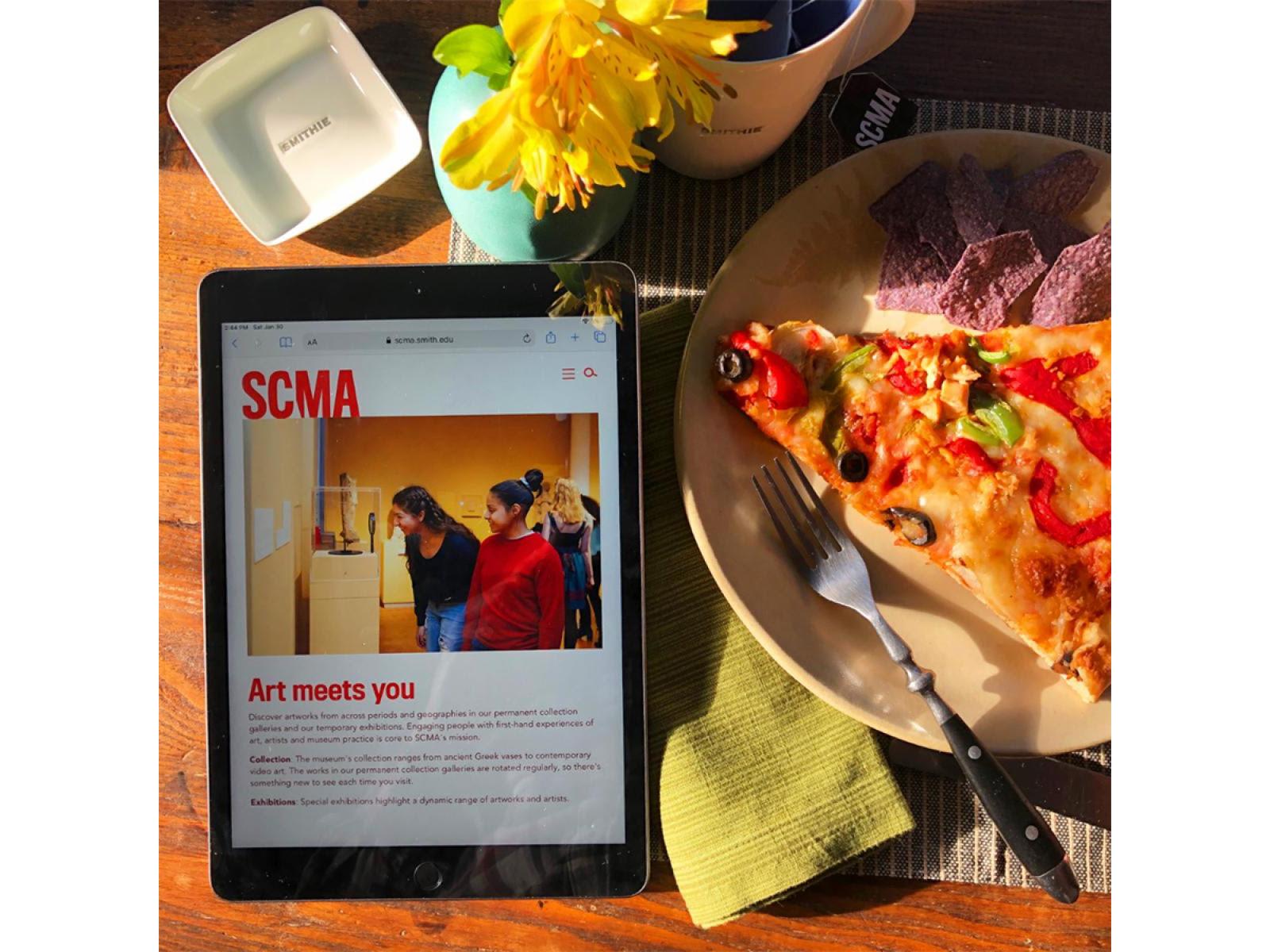 ipad with picture of SCMA website with two students looking at art. ipad on a table with pizza slice and flowers