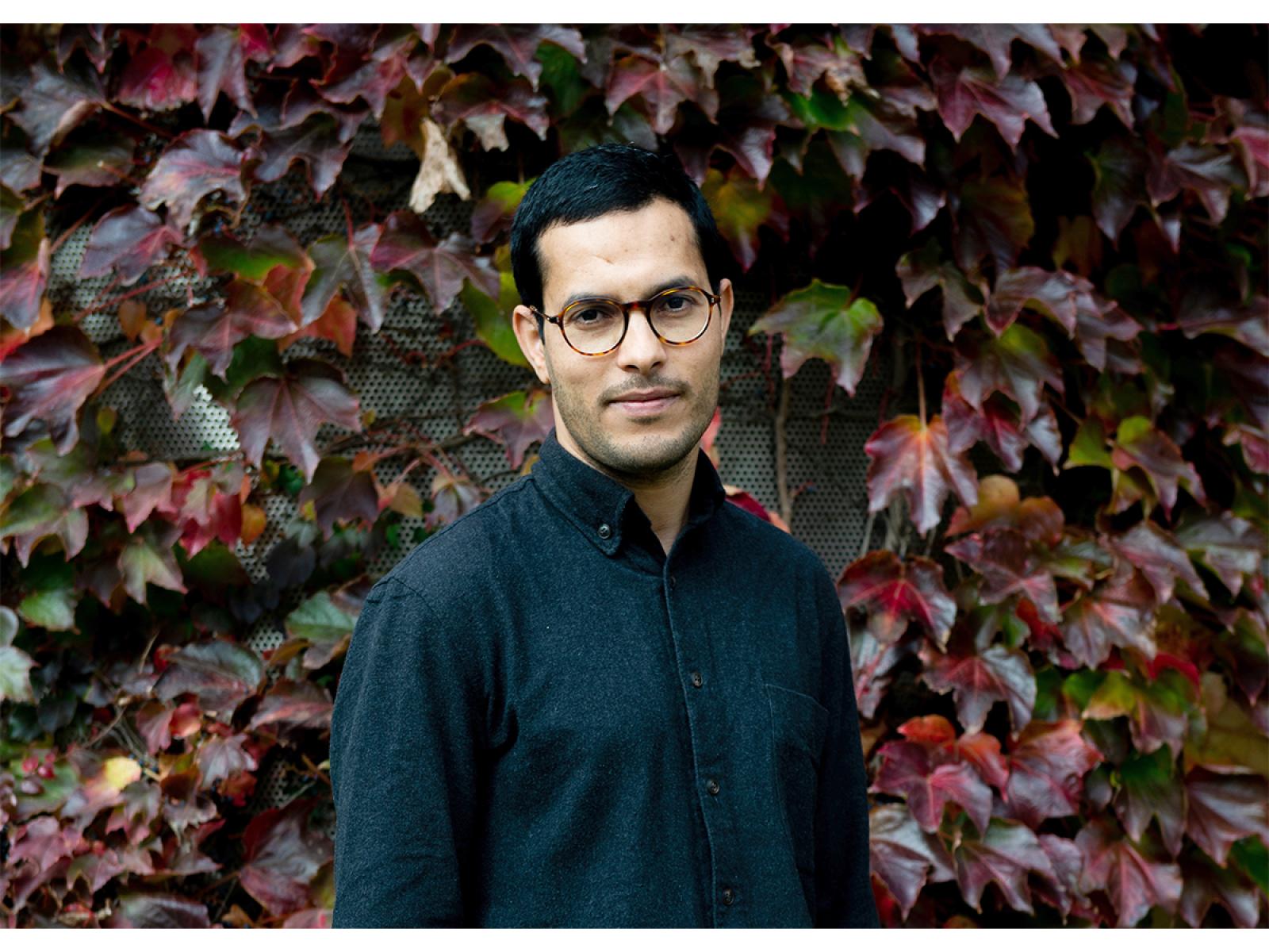 Photograph of artist with dark hair, teal shirt in front of a wall with dark red and green ivy-like plants