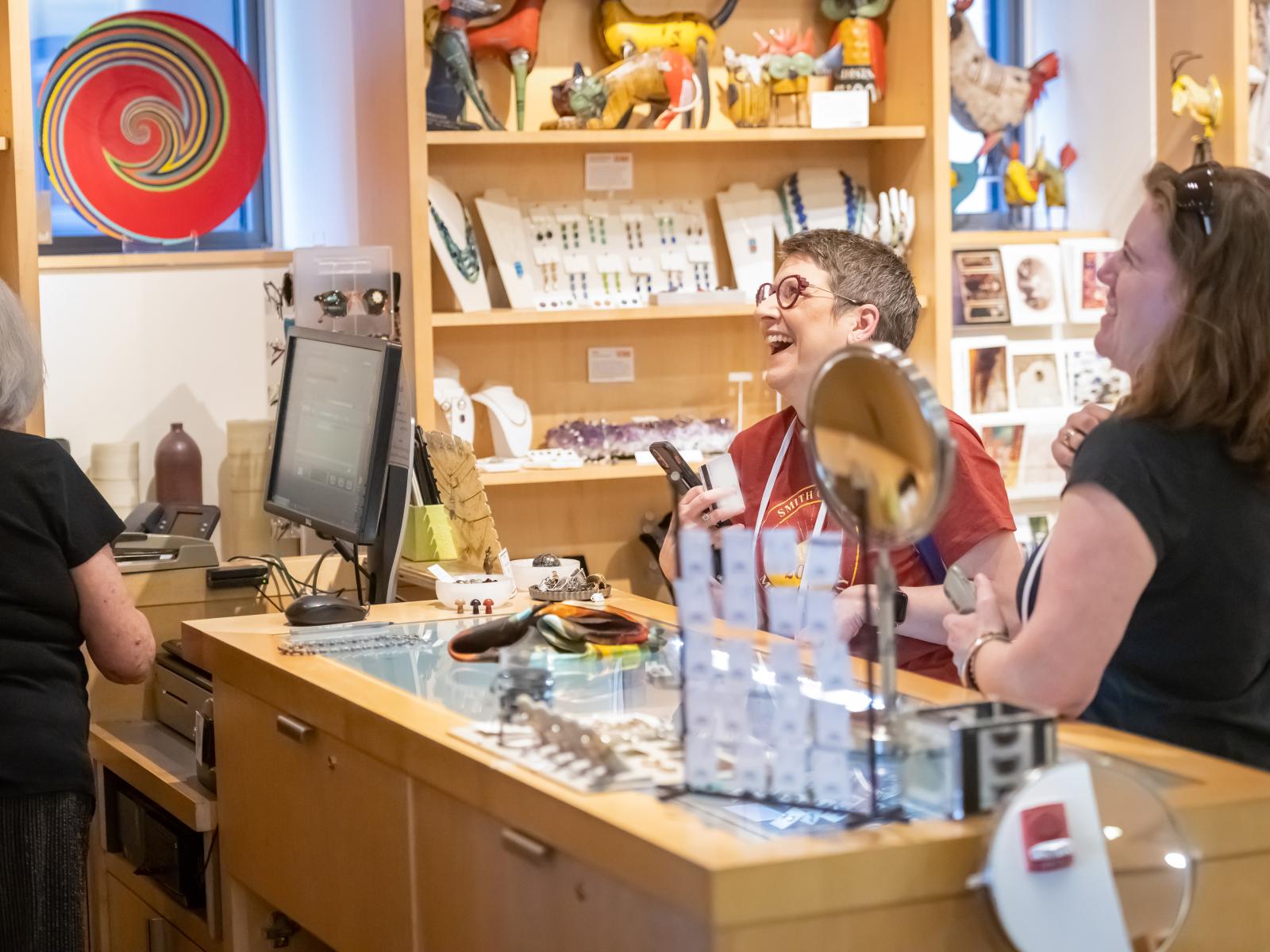 Two women smiling and laughing while standing at a wooden counter in a room full of colorful jewelry with woman in black shirt on the left of the photo