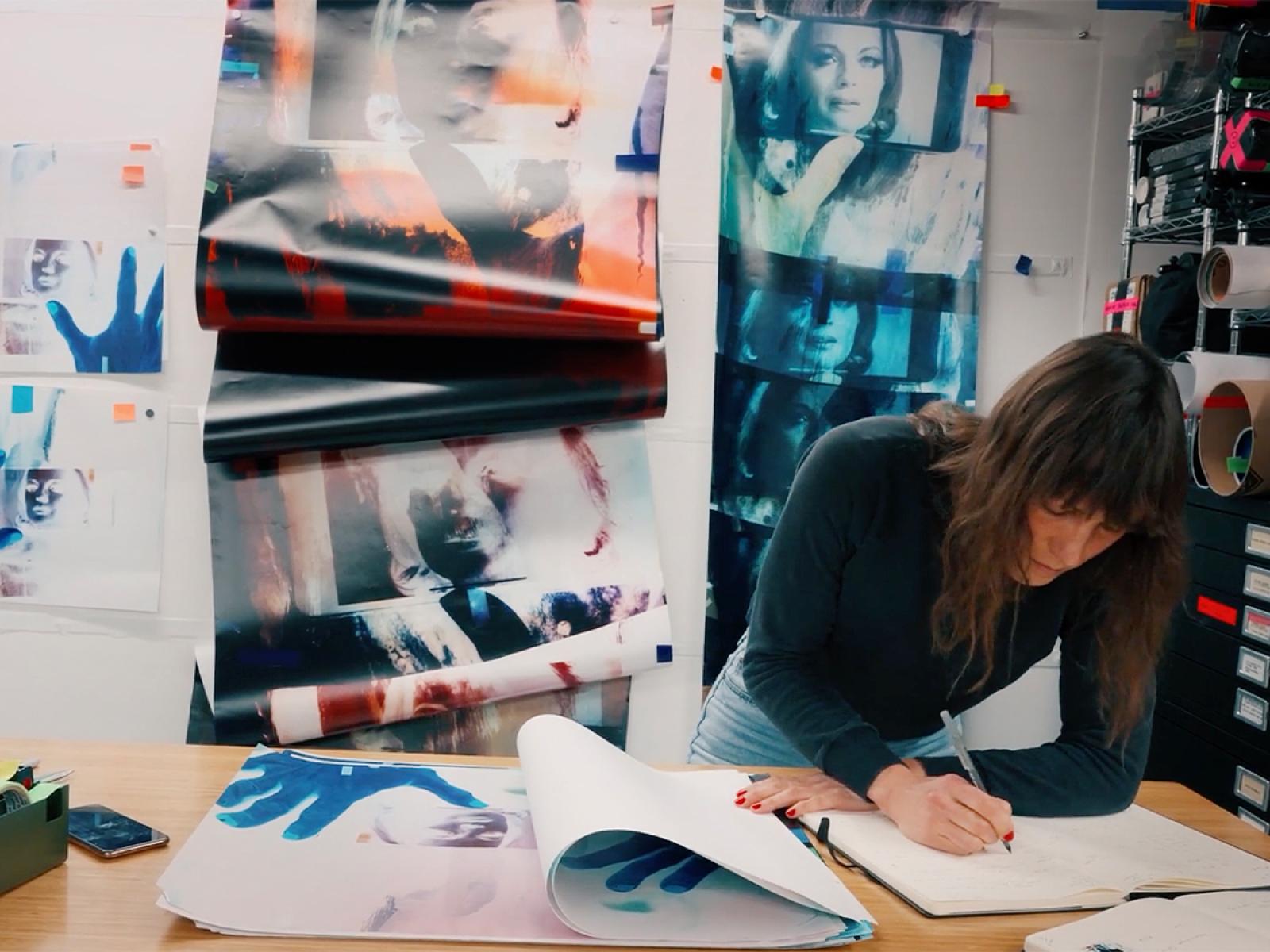 Person leaning over a table working on paper with artwork on paper all around them.