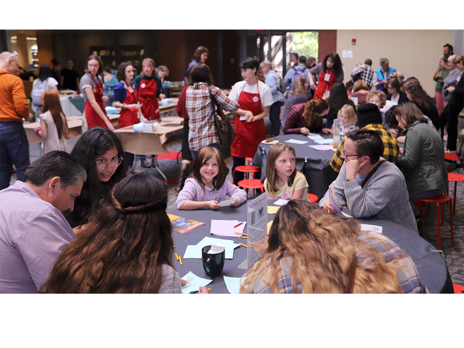 Photo of a high ceilinged room with dozens of people of varying ages engaging with art and each other.