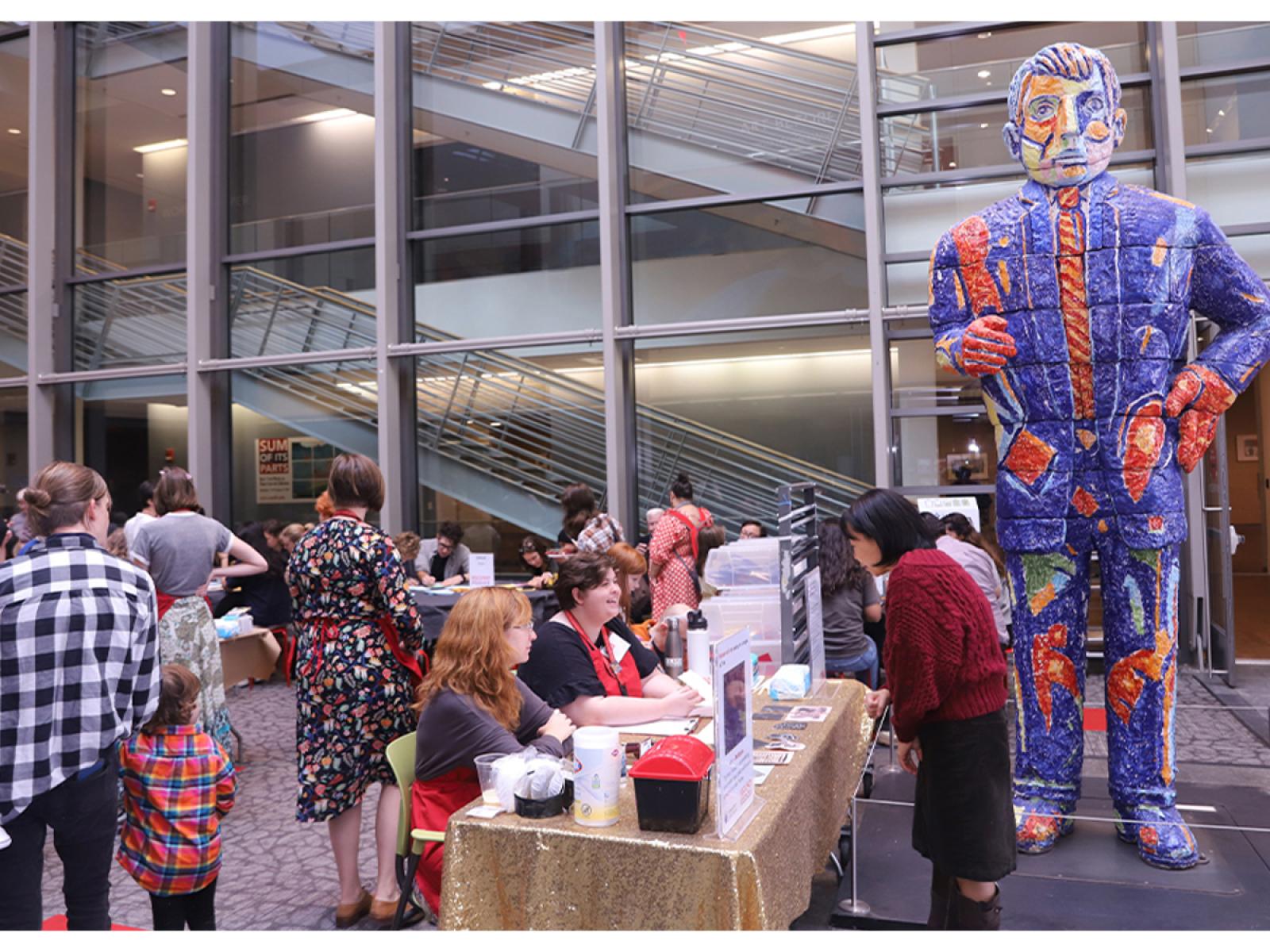 Two people sitting at a table greeting a visitor in a room with lots of people of all ages making art. 