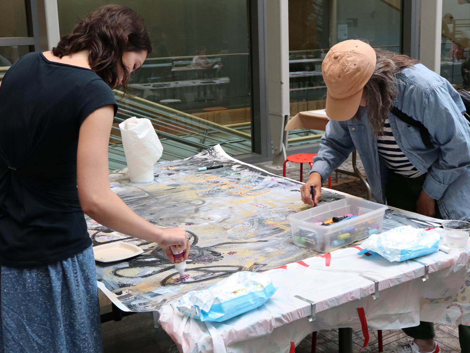 Two standing visitors working a a large art peice at the same table