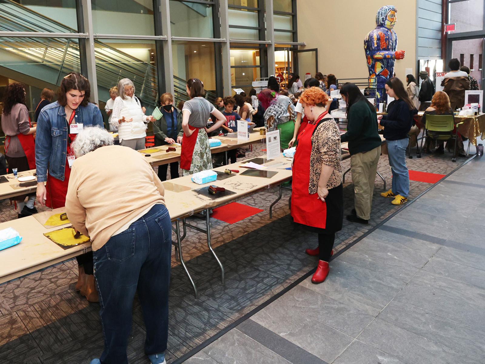 People of all ages in a large room standing and sitting while printing at long tables