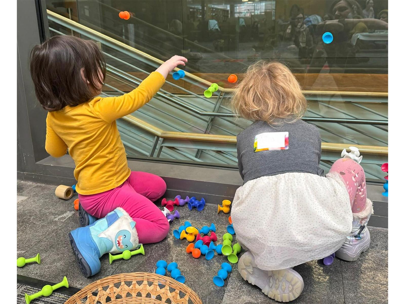 Two toddlers playing with sensory toys on a glass wall.