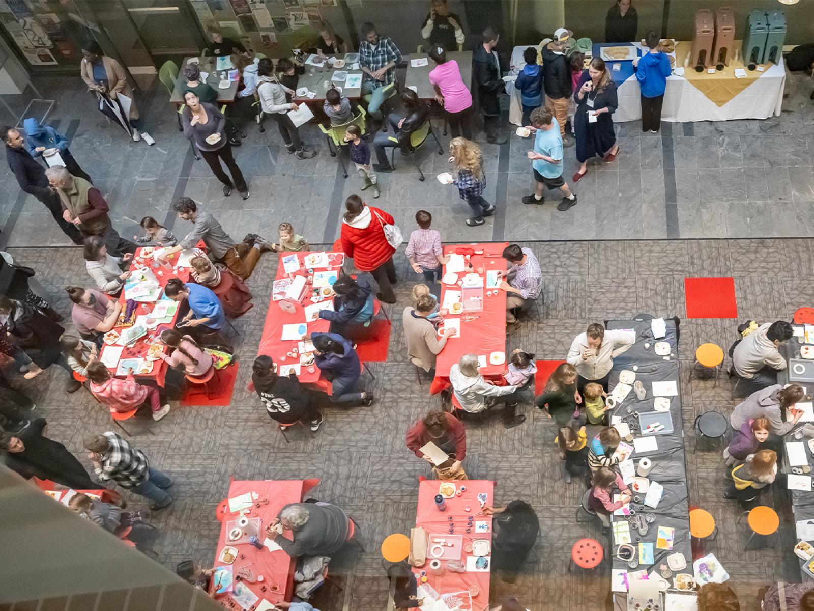 Birds Eye view of a room full of a crowd of people of all ages at tables making art and eating refreshements