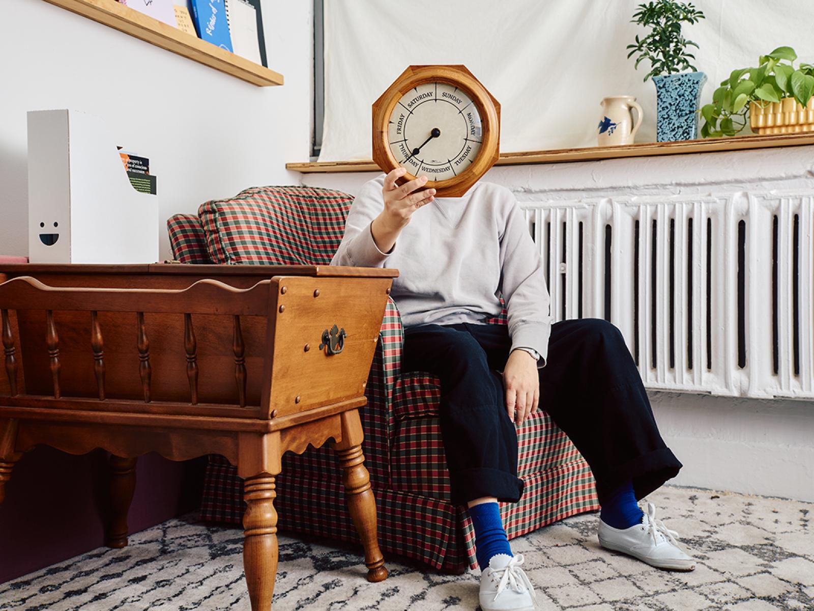 Person in room seated while holding a day of the week clock over their face. There is a table to the left