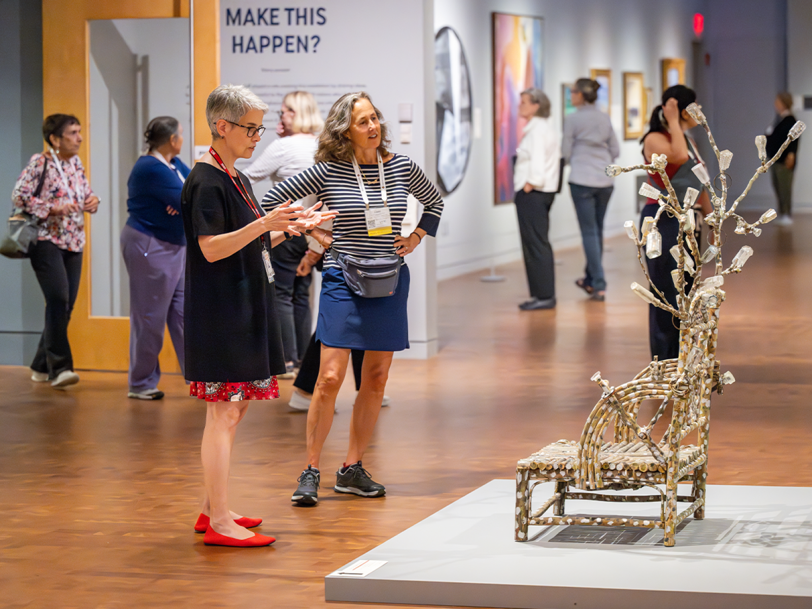 Two people in front of a chair-like sculpture in a gallery with with six people in the background 