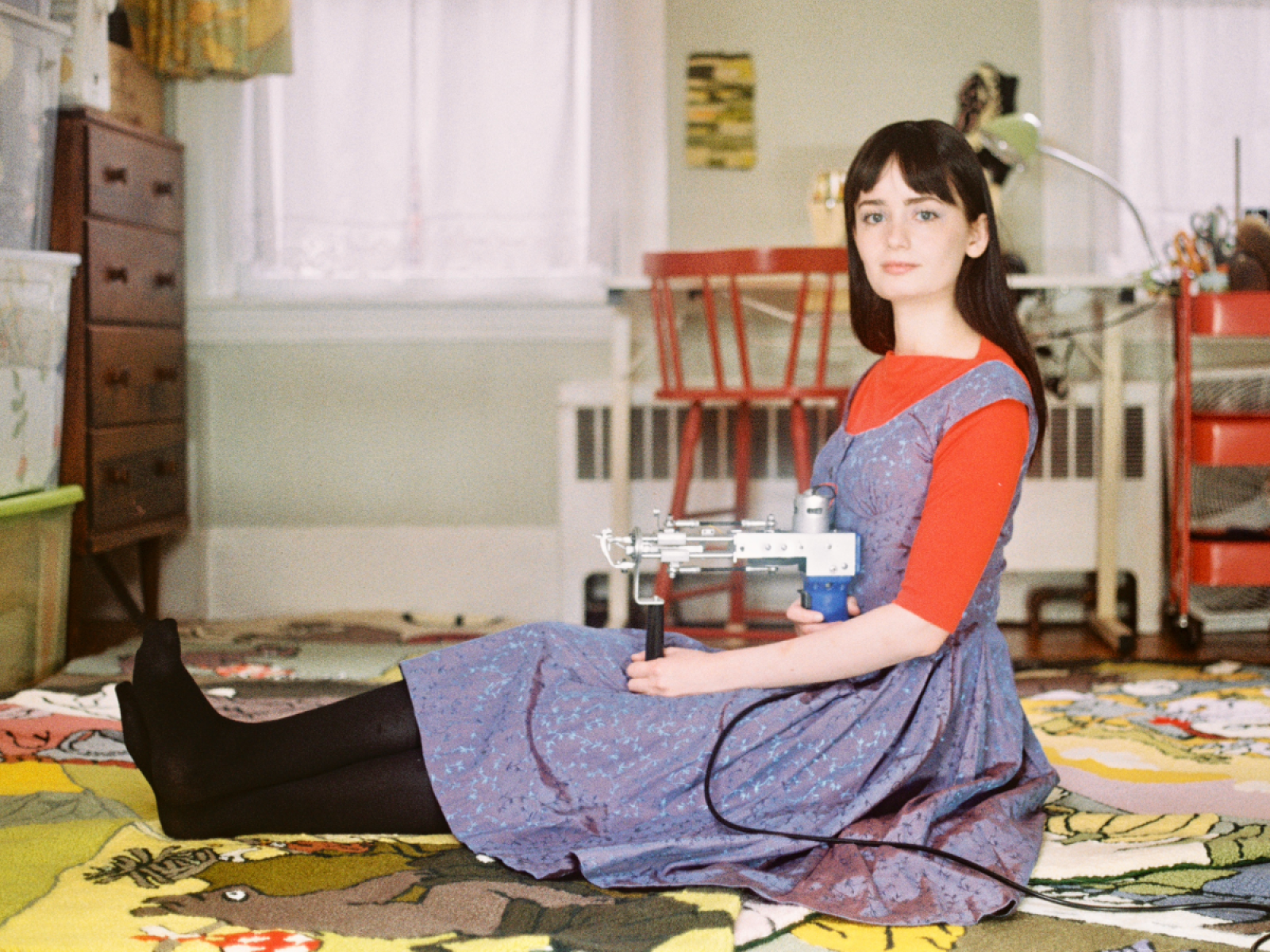 person in a denim dress sitting on a rug in a rom holding a pun needle machine on their legs