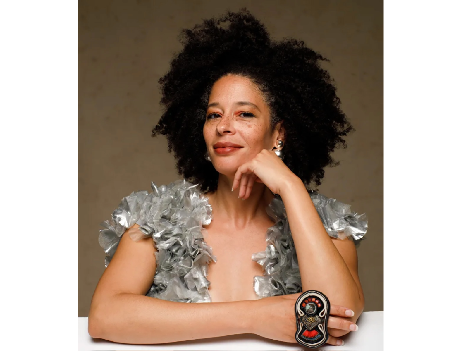 Person sitting in front of a table with curly hair, wearing a silvery dress, a large ring on hand resting on the table and has other hand resting on her cheek as they look at the viewer