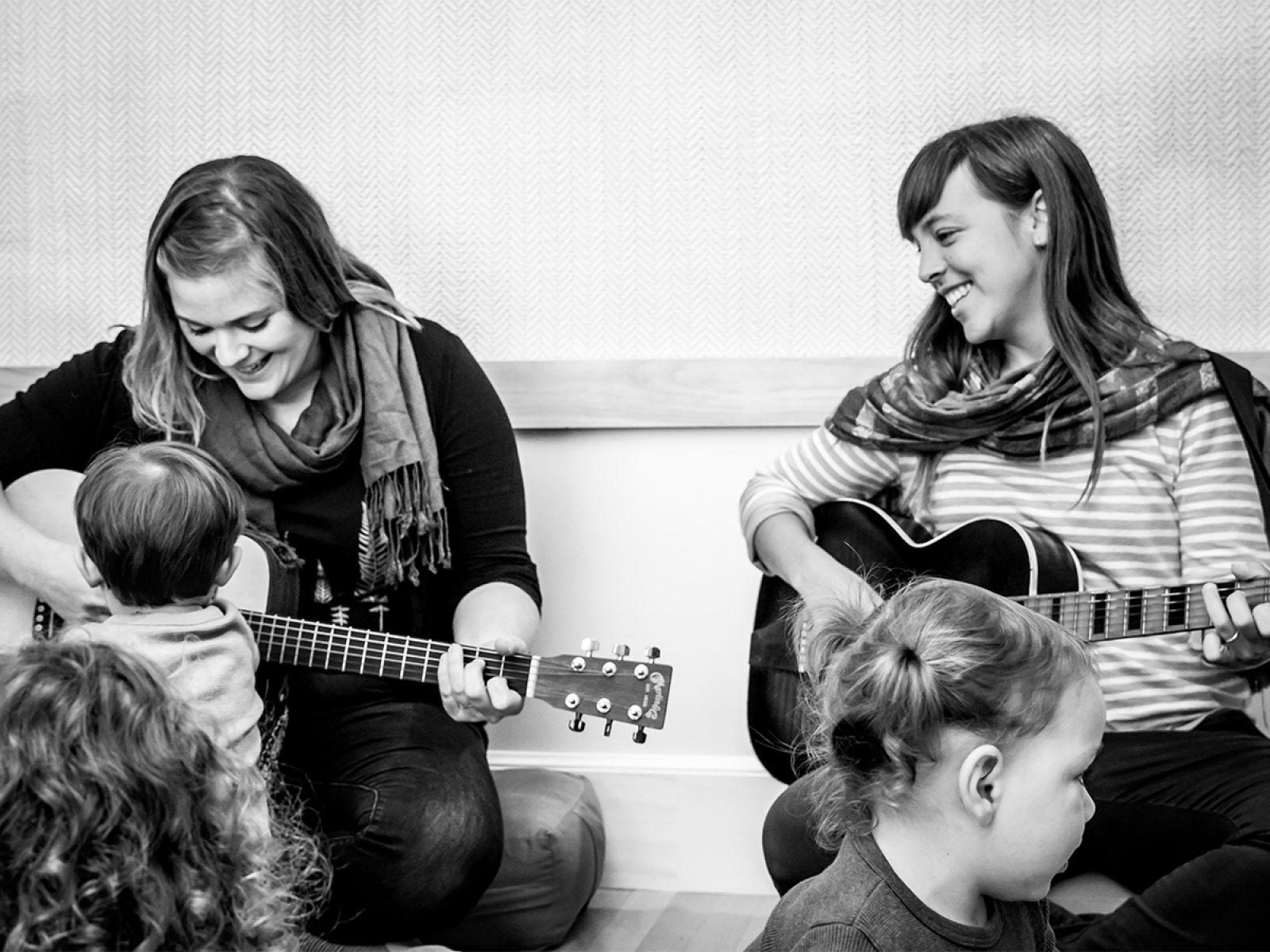 Two adults playing guitar with three young children listening and engaging with the musicians