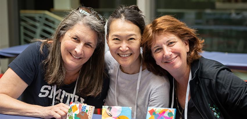 Three women posing at a table with little paintings on them.