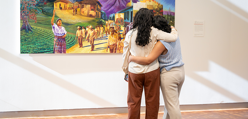 Two people with arms around each other looking at a painting of many people walking along a road together