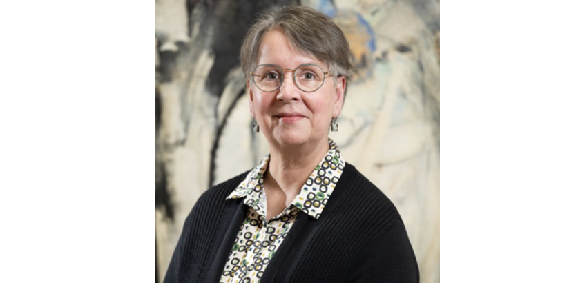 Woman from chest up in black sweater, glasses, short hair standing in front of a contemporary abstract painting with muted tones