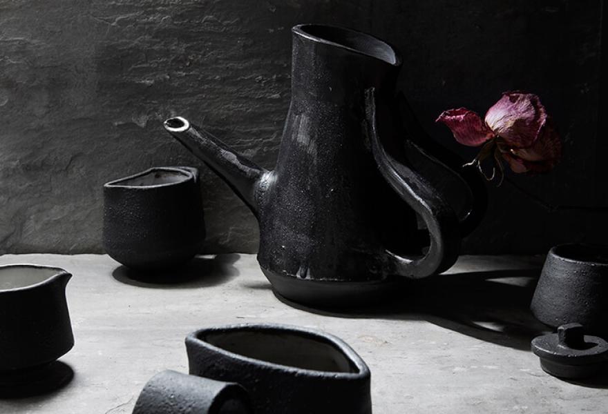 Black ceramic tea set with black tea pot and irregular mugs and with a purple flower next to the tea pot