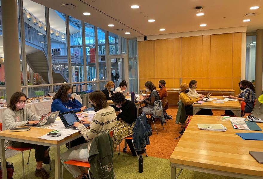 Students sitting at wodden tables in a study room with their laptops and papers. There's a glass wall looking out into an atrium with stairs