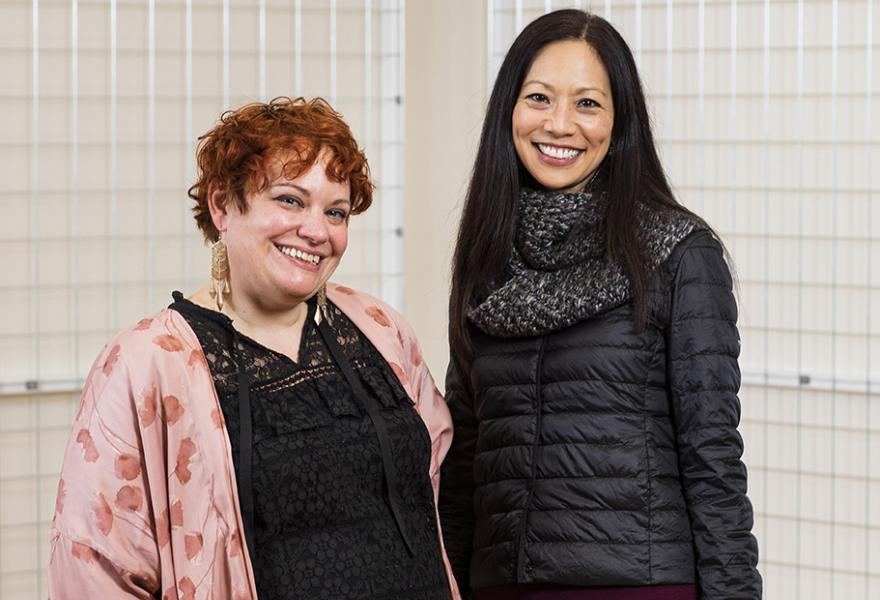 Two women standing side by side looking at camera, one on left has short red hair, a black dress and pink scarf and the on on the right is a little taller with long black hair and long black dress and jacket