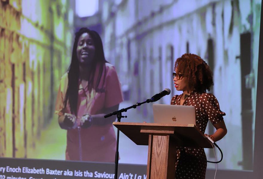 Womanwith red curly hair in dress standing at podium speaking with a projection of a person on the screen behind her