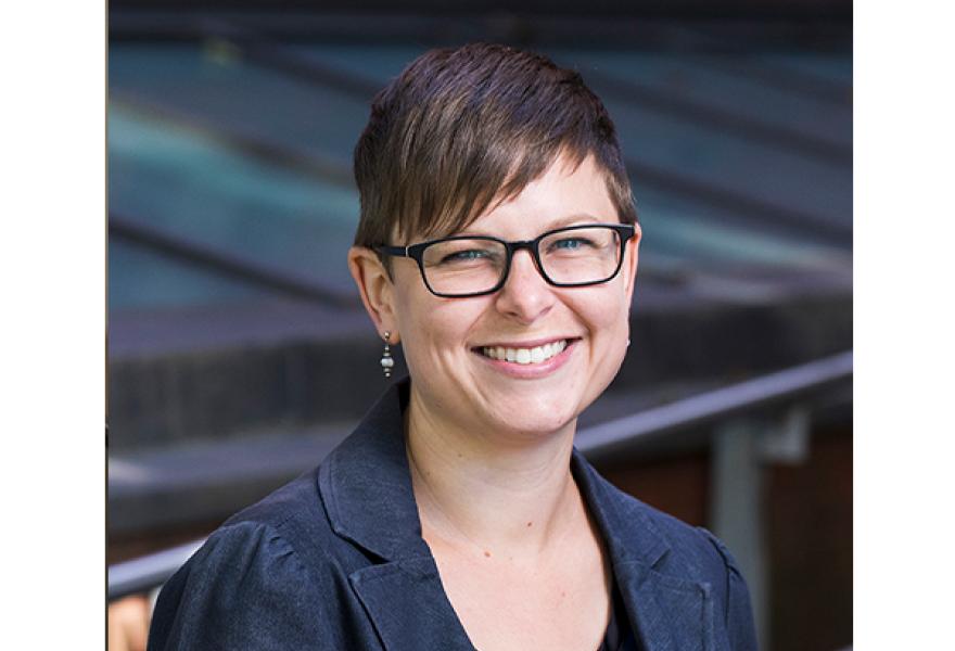 smiling person with short brown hair and glasses and blue coat jacket