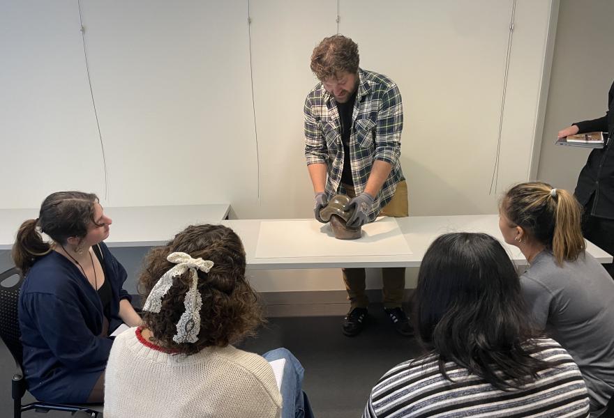 Five people sitting around a table while watching a person present a sculpture