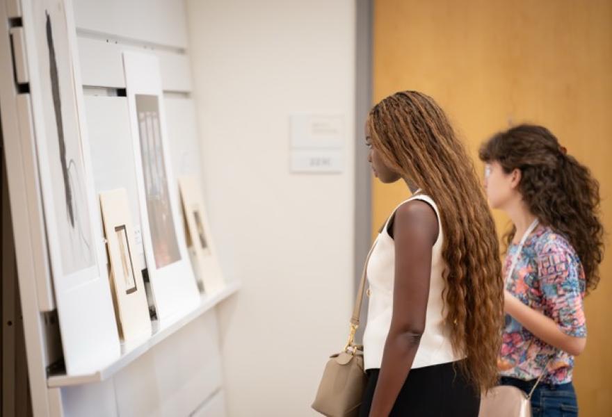 Two people on the right looking at works on paper on the wall to the left