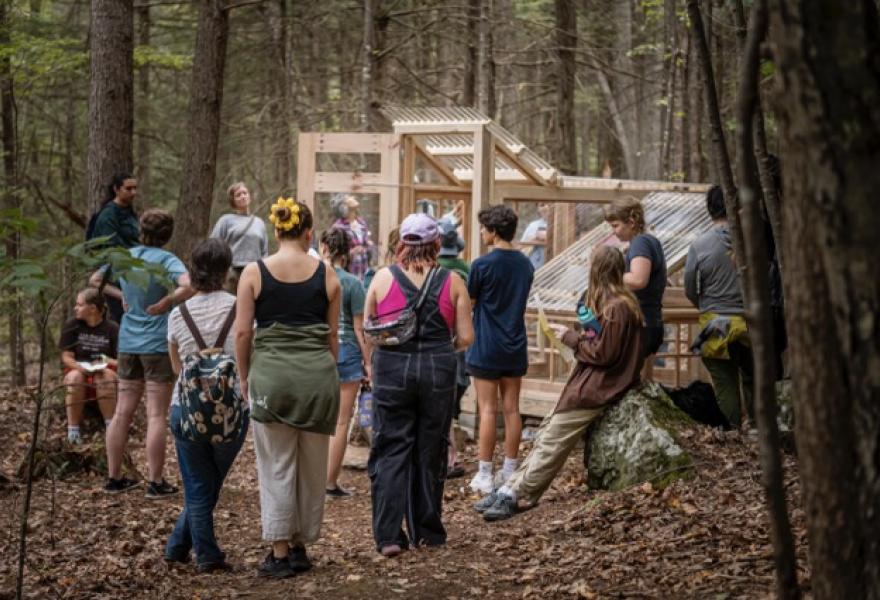 Crowd of people int he woods surrounding a wooden structure with windows and a clear plastic roof