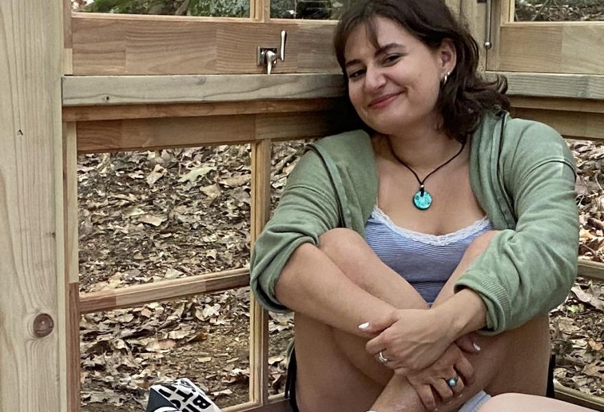 Short haired person sitting in a wooden structure with windows with legs crossed and arms holding legs