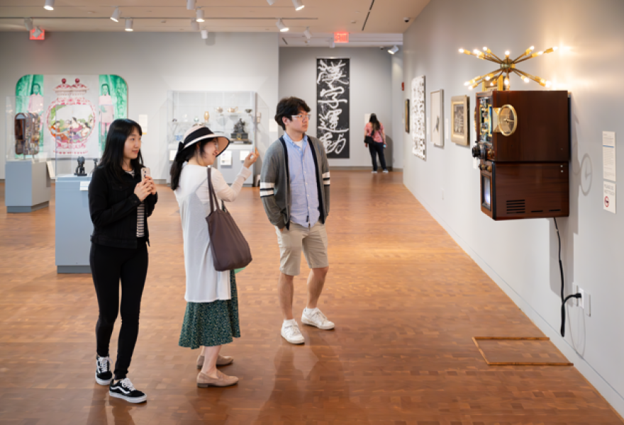Three visitors in front of an abstract wooden box sculpture with electronic screens on the front and a multi-pronged shape at top with lights on the tips