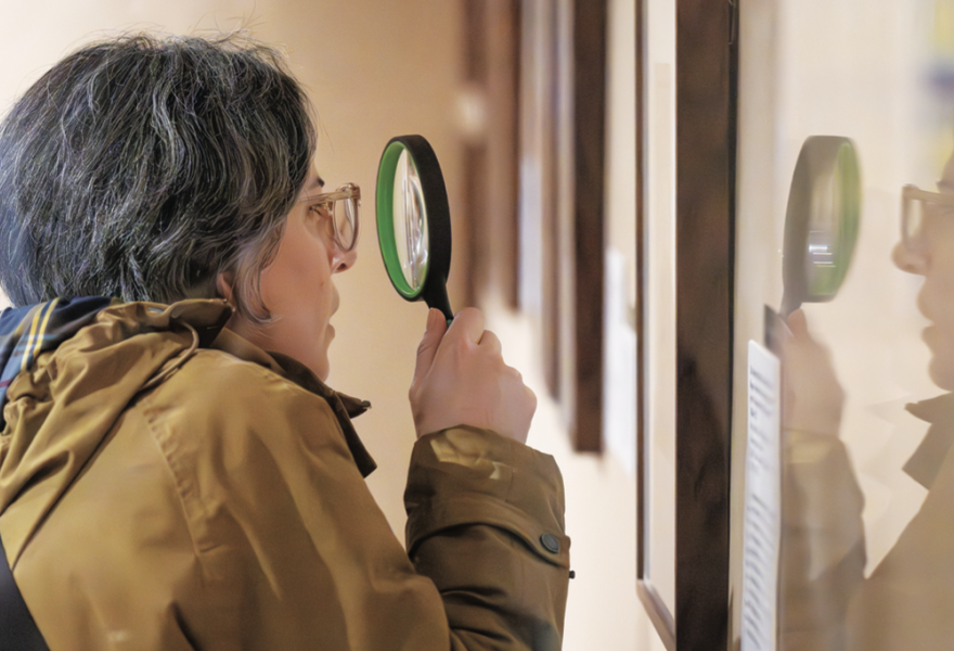 Person with graying hair and an ochre colored coat looking closely at an artwork using a magnifying glass