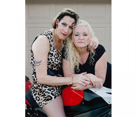 dark-haired individual in a leopard print dress seated on a motorcycle with their arms around a blond individual dressed in black