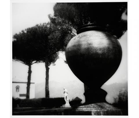 close-up black and white image of an urn in the foreground, with a white figural statue at the center and trees in the background