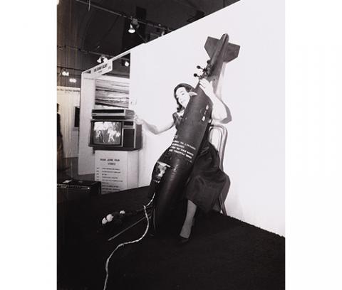 black and white image of woman playing a cello shaped like a bomb in front of a white wall with a video monitor to her right